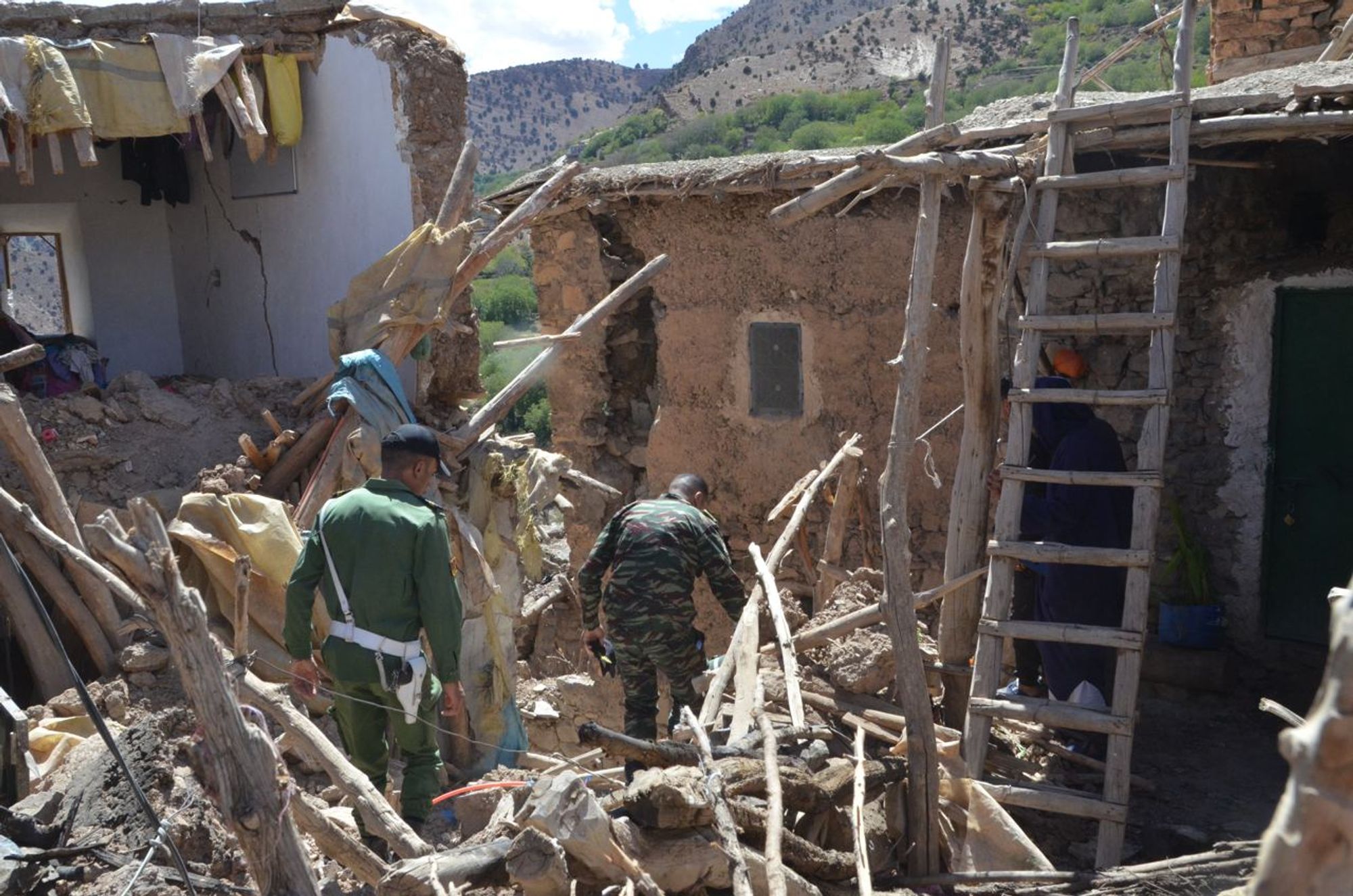 Two military personnel amid walking through rubble and partially collapsed house.
